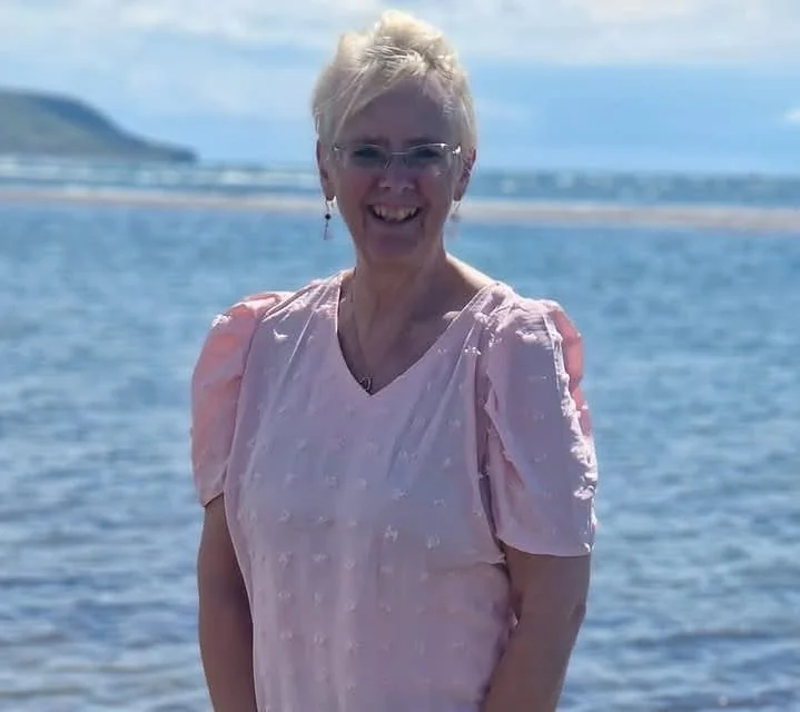 Smiling woman with short blonde hair and glasses standing on a beach with ocean and sky in the background.