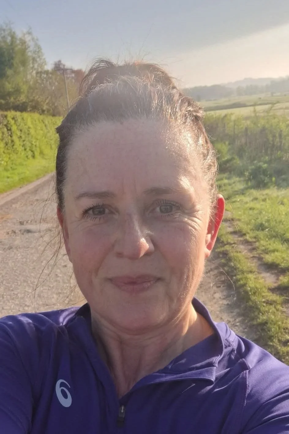 A woman taking a selfie outdoors during sunset, with green fields and a dirt path in the background.