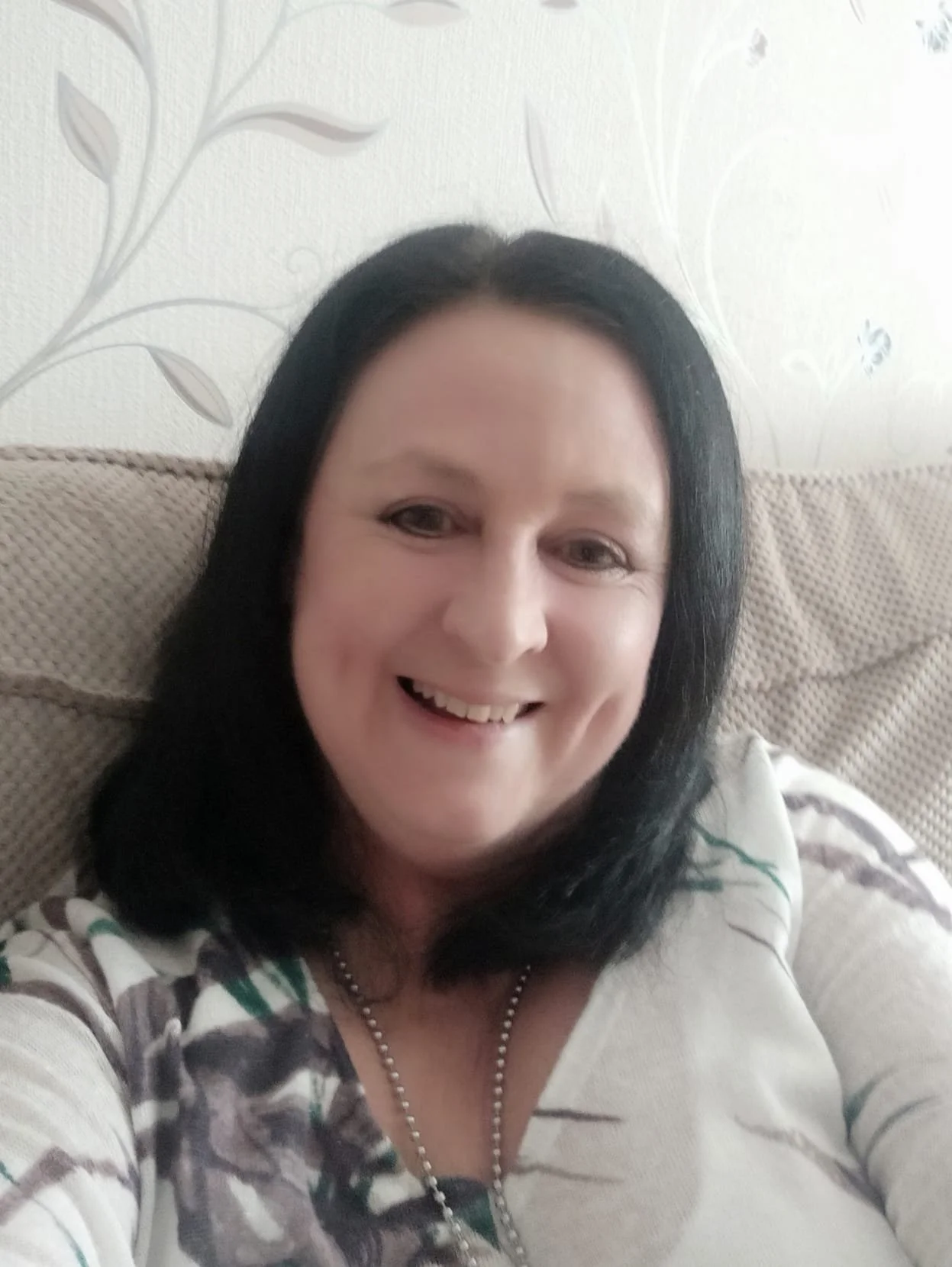 A woman with black hair smiling and taking a selfie, sitting on a beige couch with a light-colored background with floral patterns.
