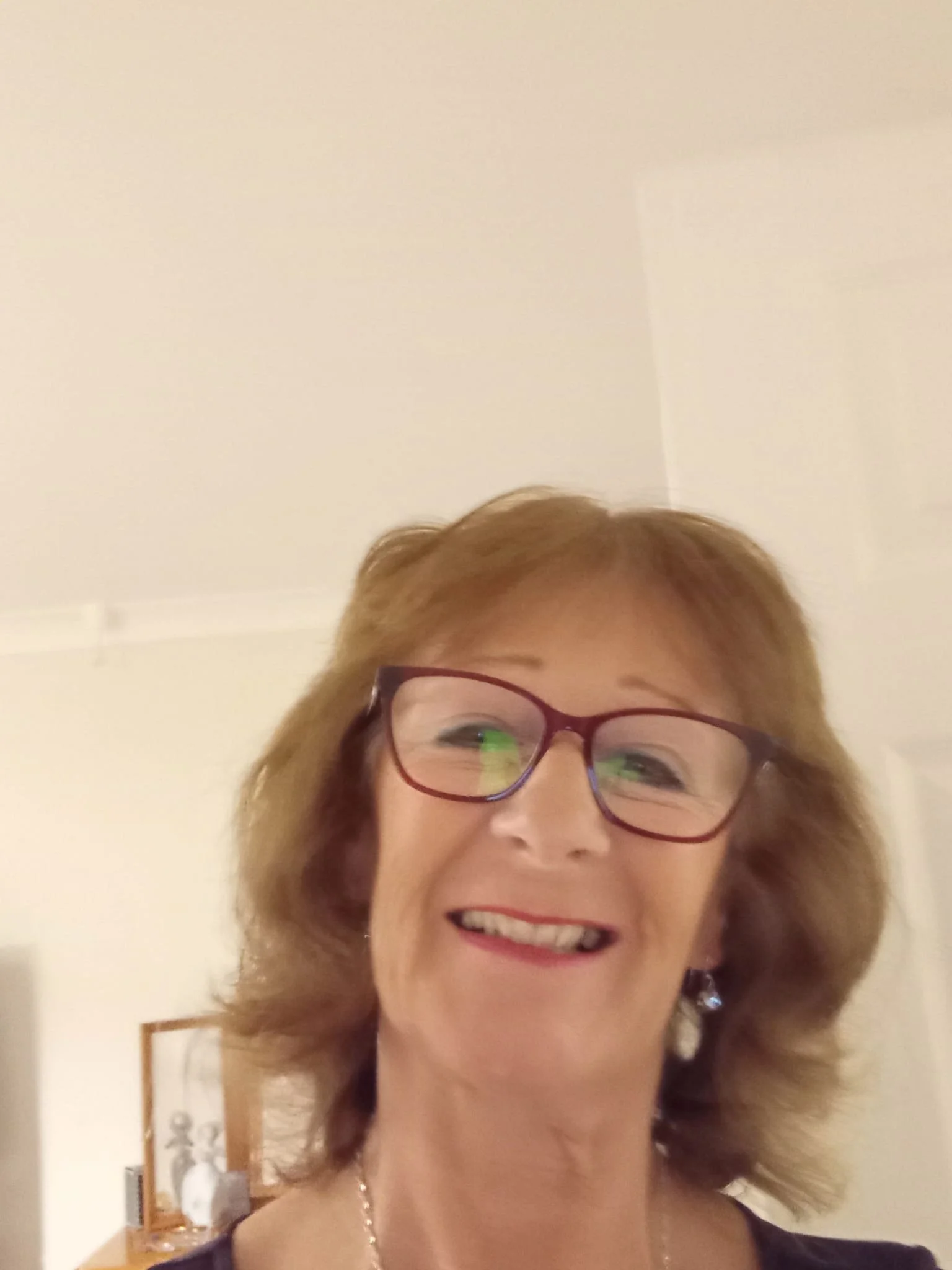 A smiling middle-aged woman with glasses, short blond hair, and pearl earrings, standing in a room with a picture frame and decorative items behind her.