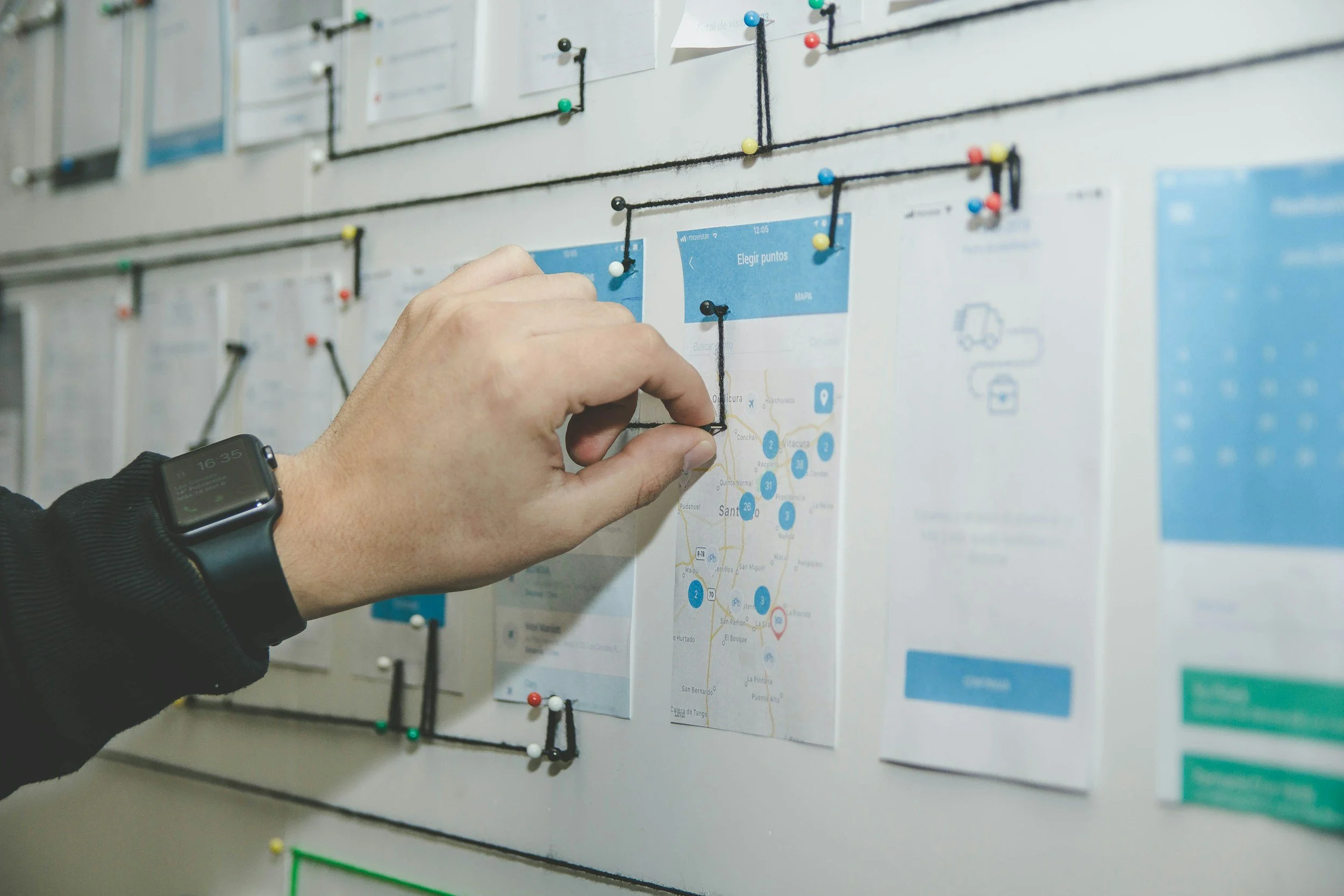 A person pinning a printed map and documents on a wall with black string and colorful pins.