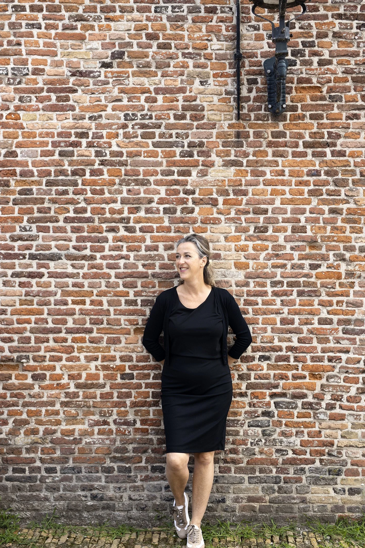 Sharon van de Veerdonk, oprichtster van weconnect.business, gekleed in een zwarte jurk en sneakers, staand voor een bakstenen muur.
