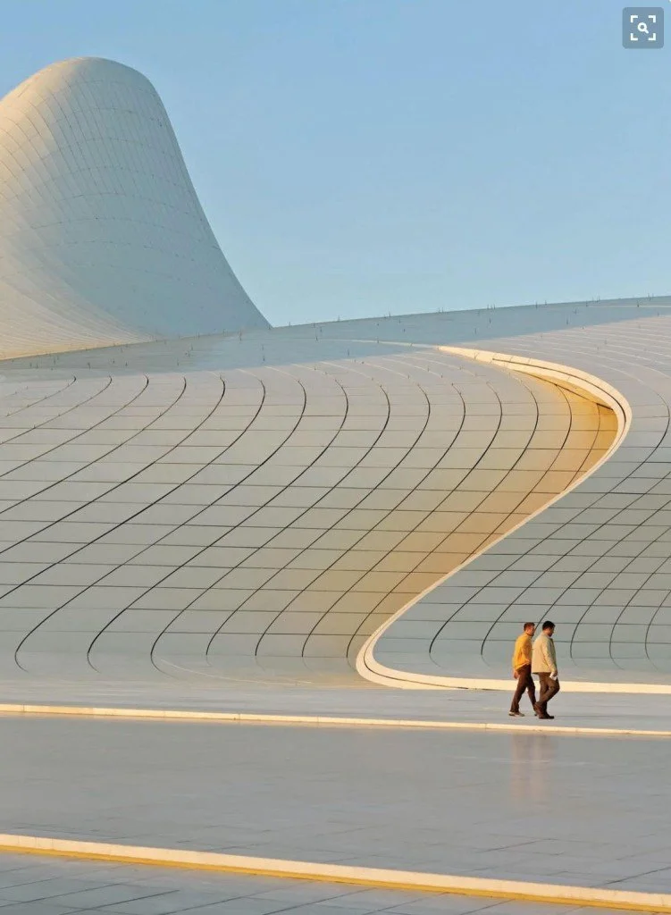 Deux personnes marchant devant une structure architecturale moderne avec des courbes et des surfaces lisses, sous un ciel bleu clair.