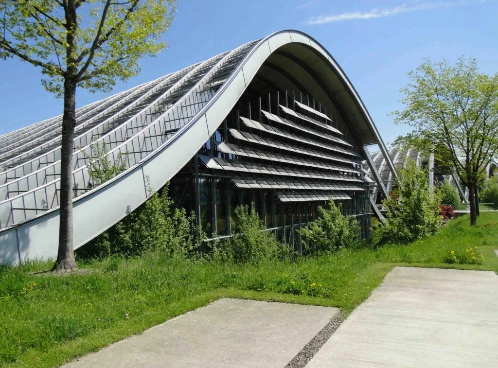 Bâtiment moderne avec toiture incurvée et panneaux solaires, entouré d'arbres et de verdure.