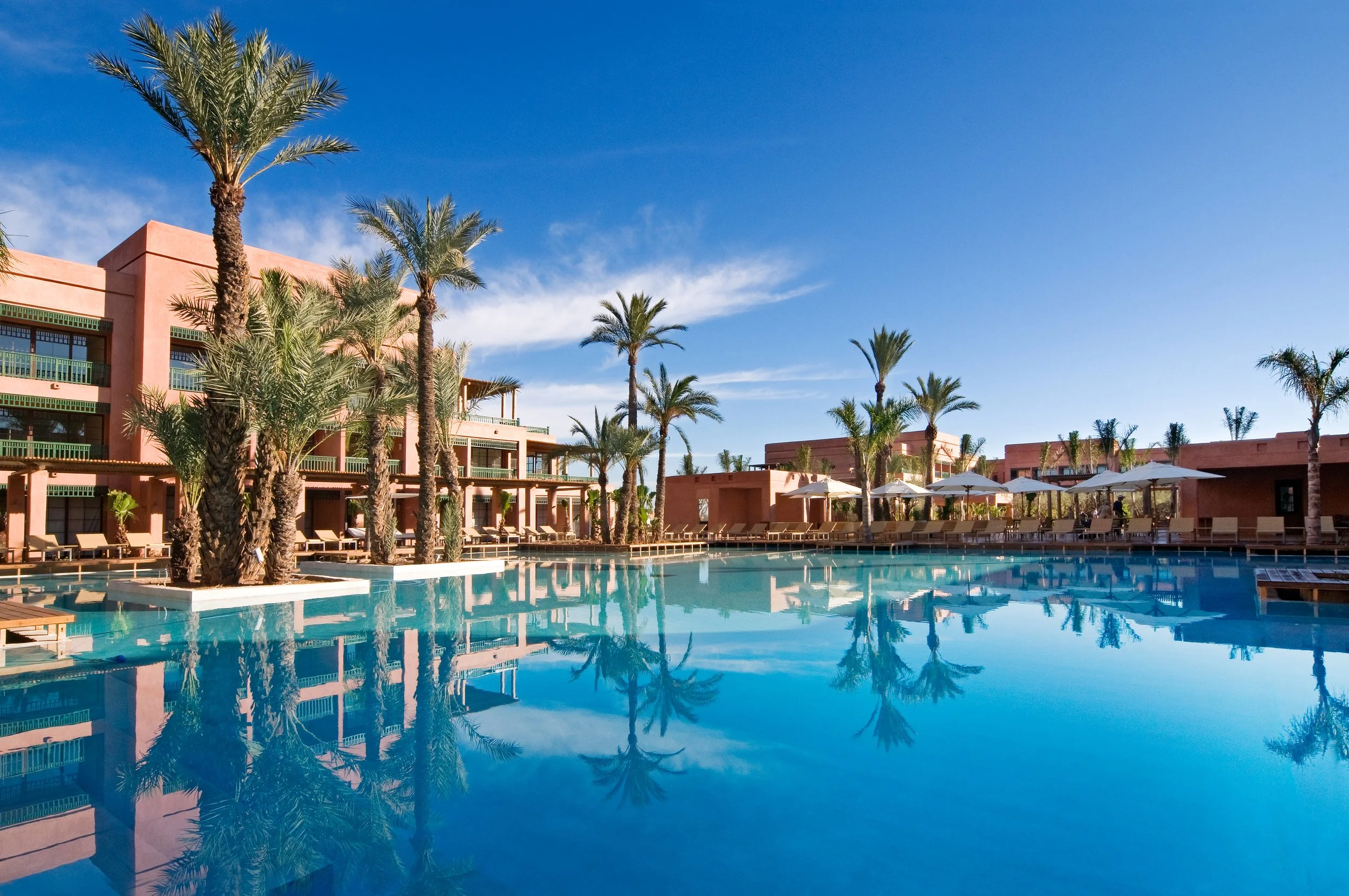 Piscine extérieure dans une station balnéaire avec des palmiers, des parasols et des transats, bâtiments en style marocain en arrière-plan, ciel bleu clair.