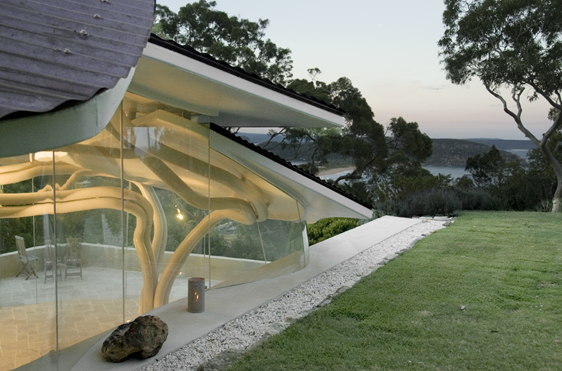 Maison moderne avec grande baie vitrée offrant une vue panoramique sur un paysage naturel, y compris des arbres et un plan d'eau. L'architecture est intégrée à l'environnement, avec des éléments en bois apparents et un toit incliné.