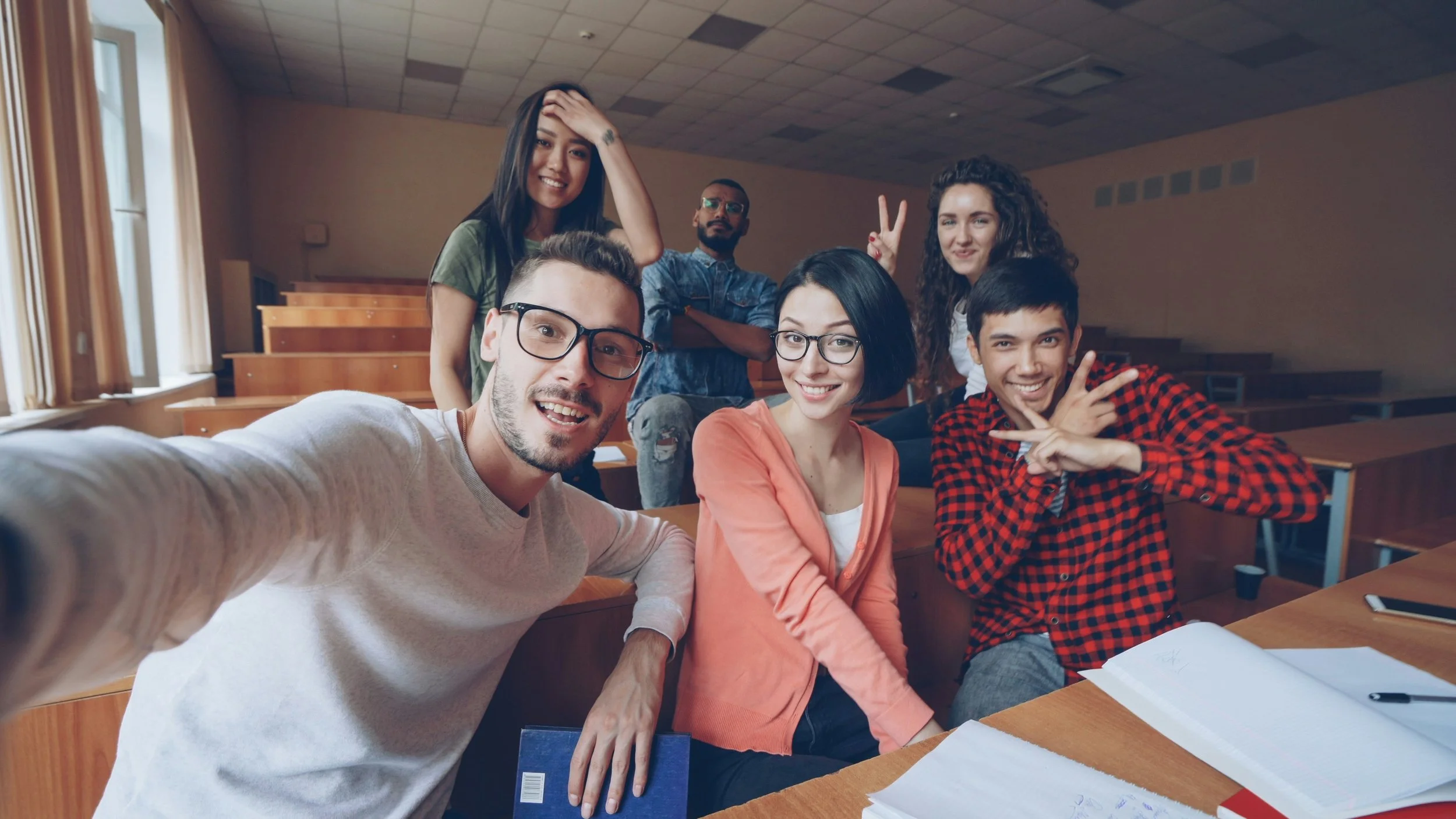 A group of six college students posing playfully for a group selfie inside a university lecture hall with tiered wooden desks.