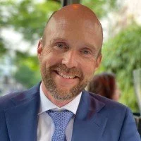 A smiling man with a beard in a suit, outdoors with greenery in the background.