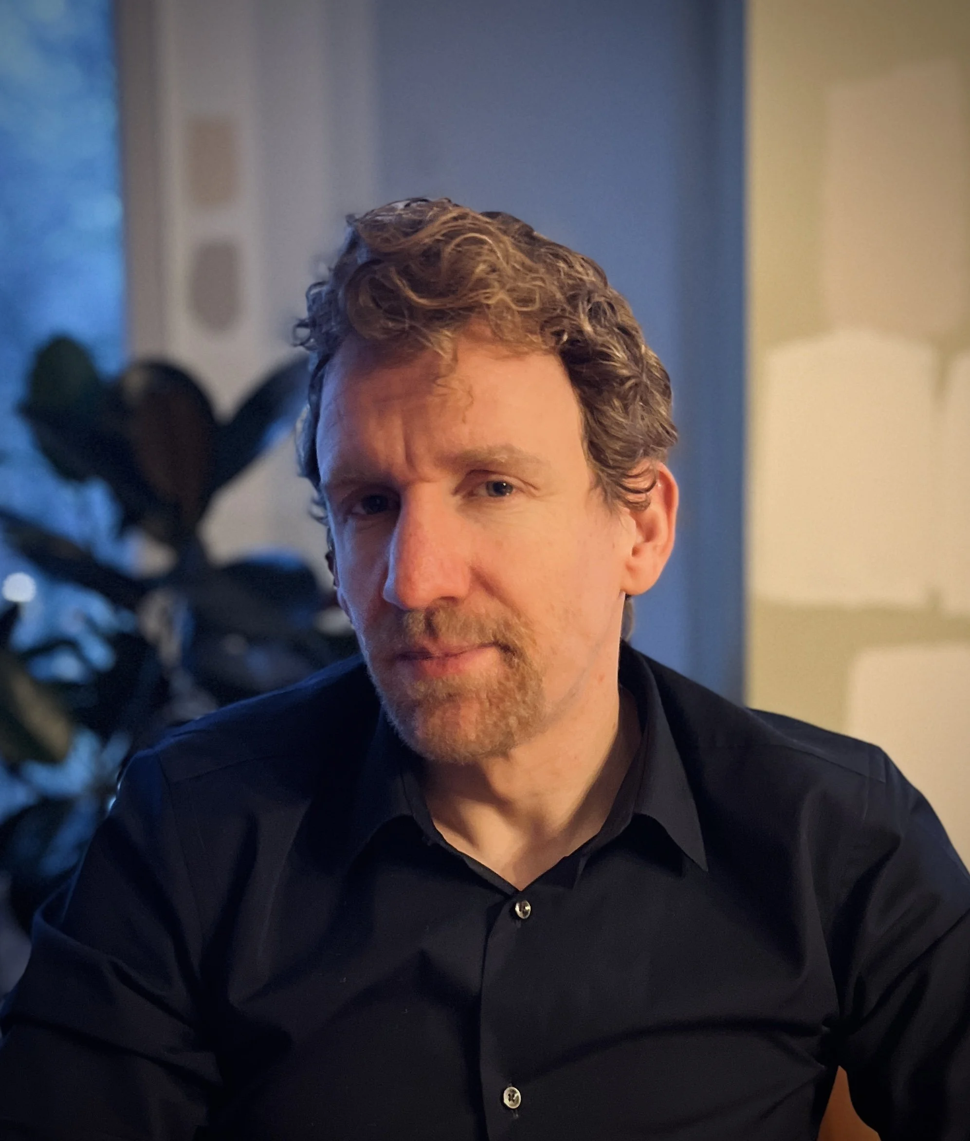 A man with curly hair and a beard wearing a black shirt sitting indoors with a plant and a lamp in the background.