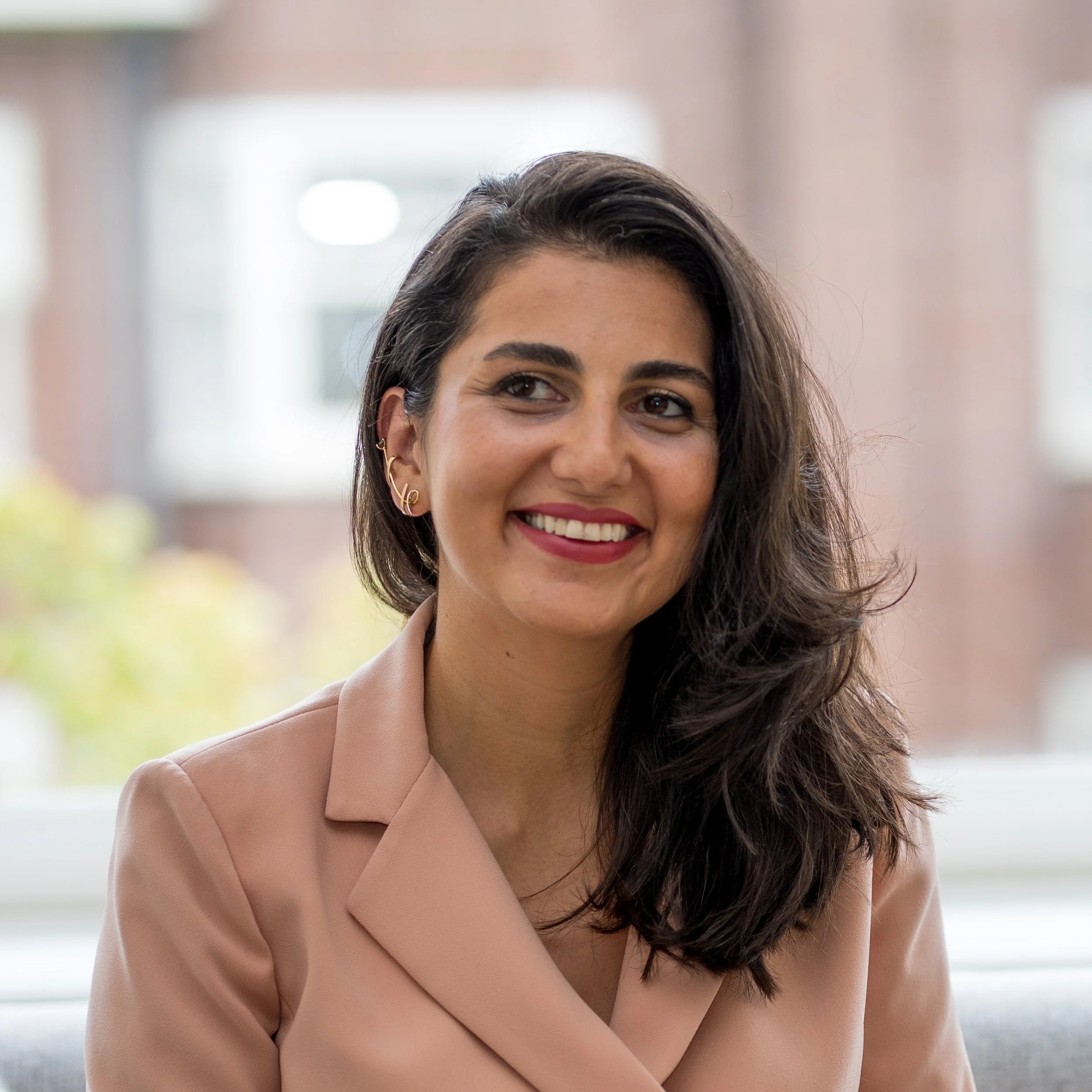 A woman with dark hair, gold earrings, and pink lipstick, smiling while sitting indoors near a window.