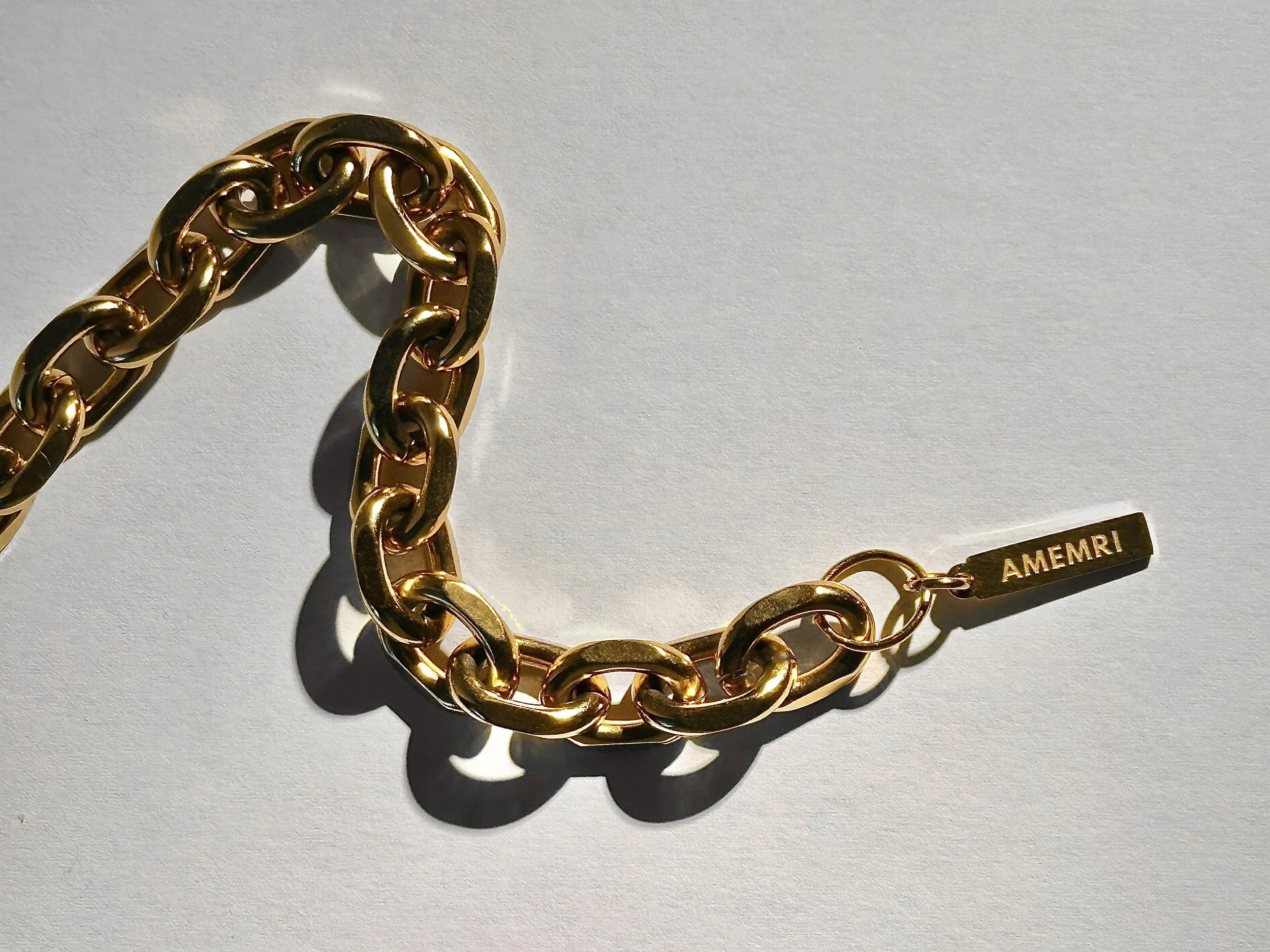 A gold chain necklace with large, interlocking links, a tag with the brand name 'AMEMRI'