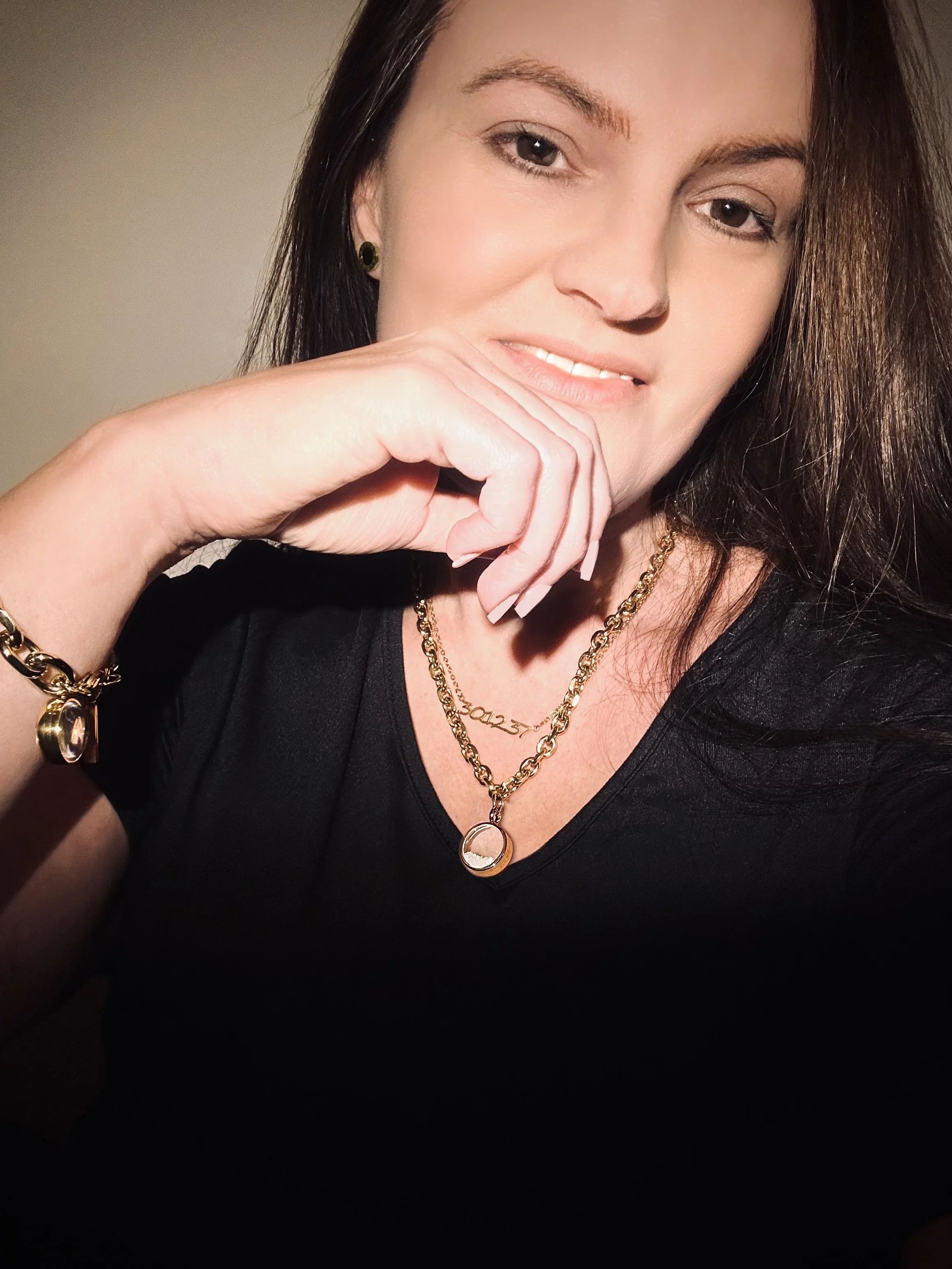 A woman with dark hair, wearing gold jewelry, and a black top, smiling and resting her chin on her hand.