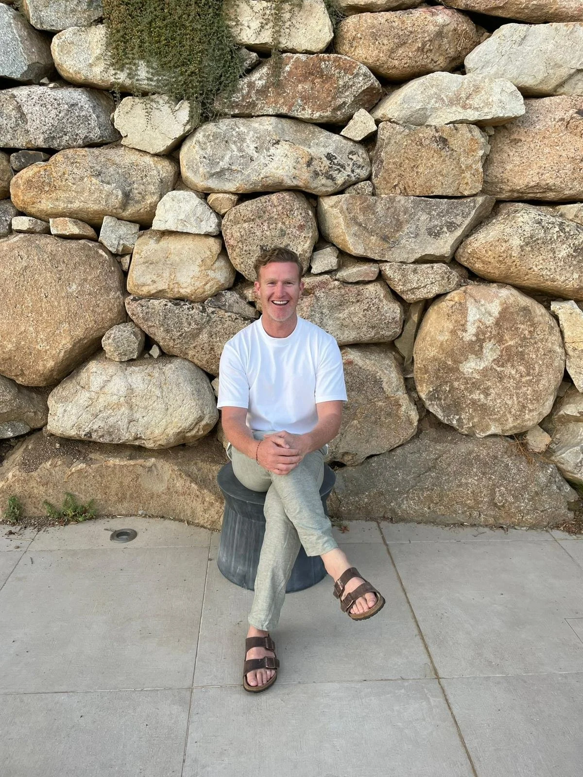A smiling man in a white t-shirt and beige pants sitting on a small stool in front of a large stone wall. He has short hair and is wearing sandals. His hands are clasped in his lap and he appears relaxed and happy.
