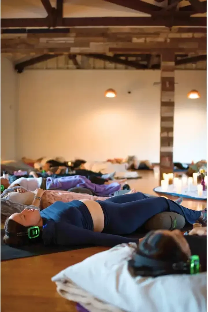 Group of people lying on mats or blankets, listening to a woman with headphones during a meditation or relaxation session in a spacious room with wooden beams and candles.
