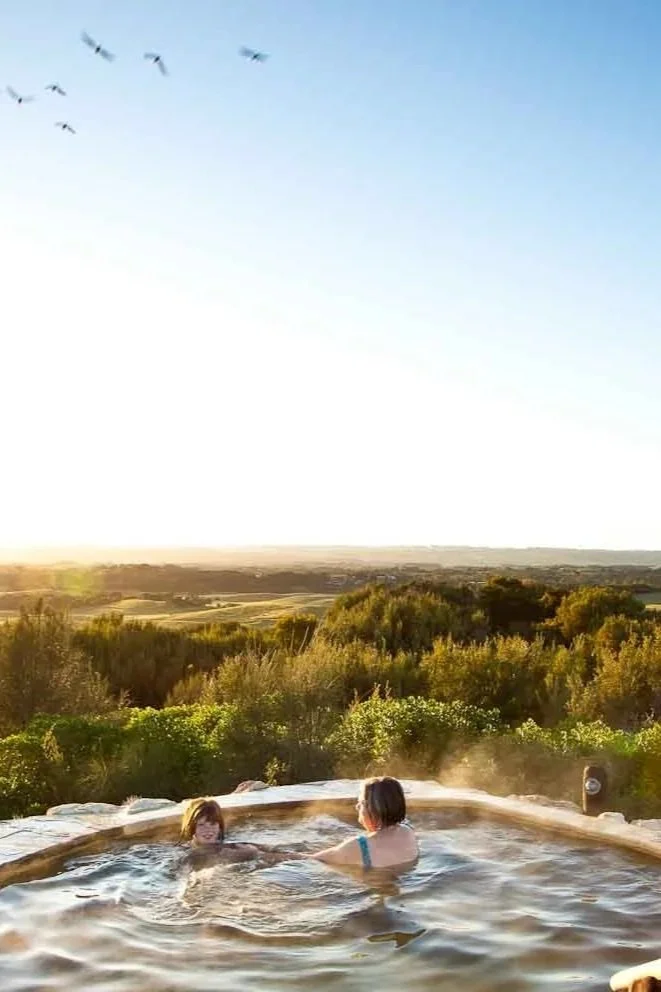 Two women sitting in an outdoor hot tub with a scenic view of trees, fields, and a clear blue sky at sunset or sunrise. Birds are flying overhead.