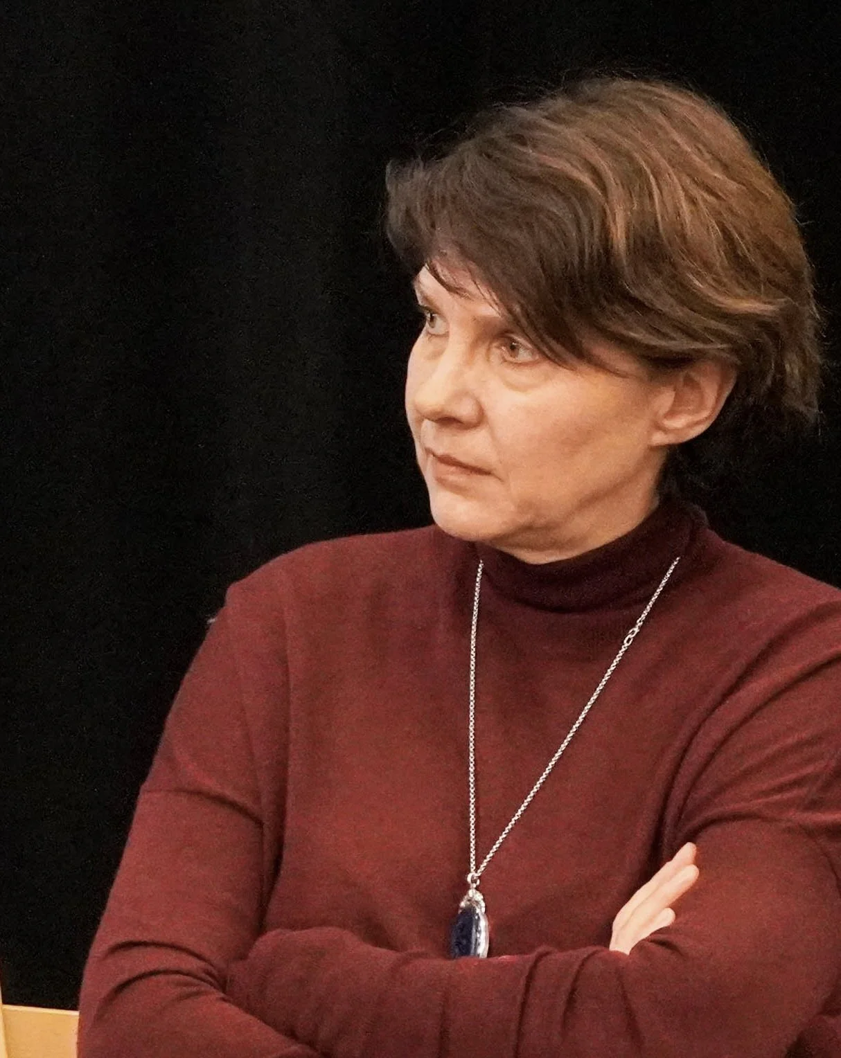 Frau in rotem Rollkragenpullover mit Schmuck sitzt nachdenklich mit verschränkten Armen vor schwarzem Hintergrund.