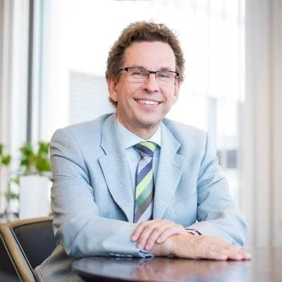 Ein lachender Mann in einem hellblauen Anzug und mit Brille, sitzend an einem Tisch in einem modernen Büro.