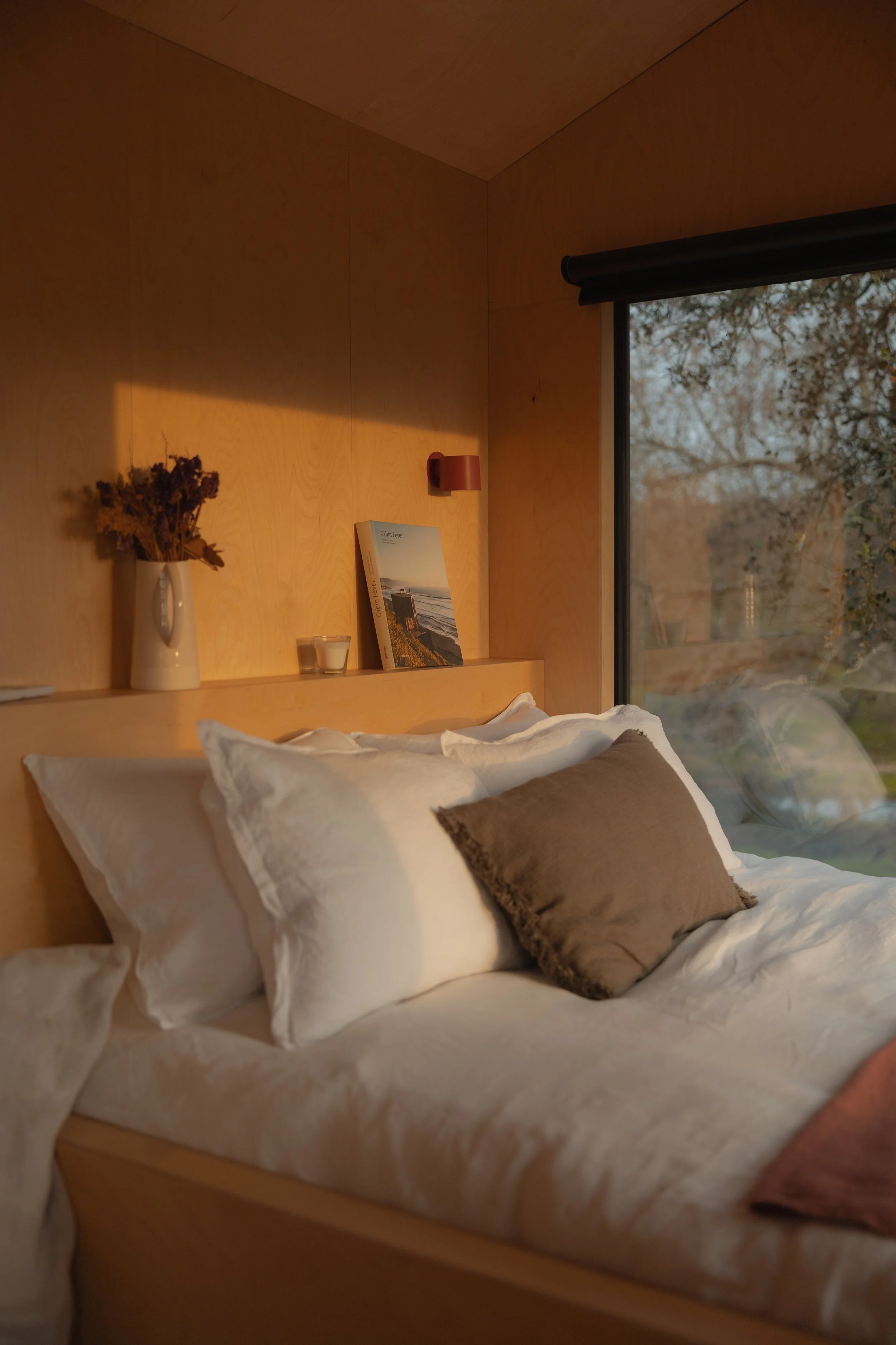 Cozy bedroom interior with bed, pillows, wooden walls, window view, and decor on a shelf with a book and vase.