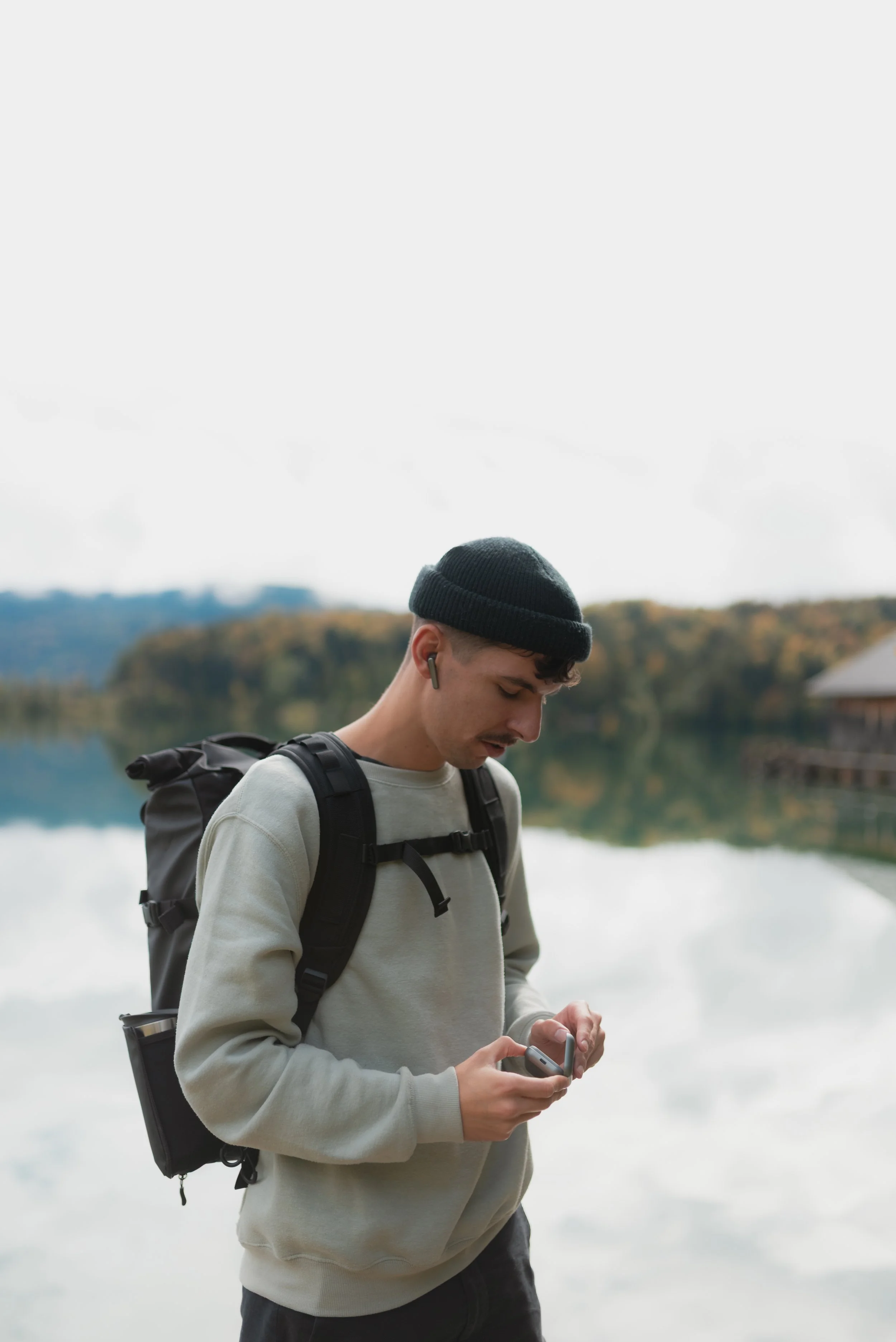 Person wearing a black beanie and backpack, holding an electronic device by a lake.
