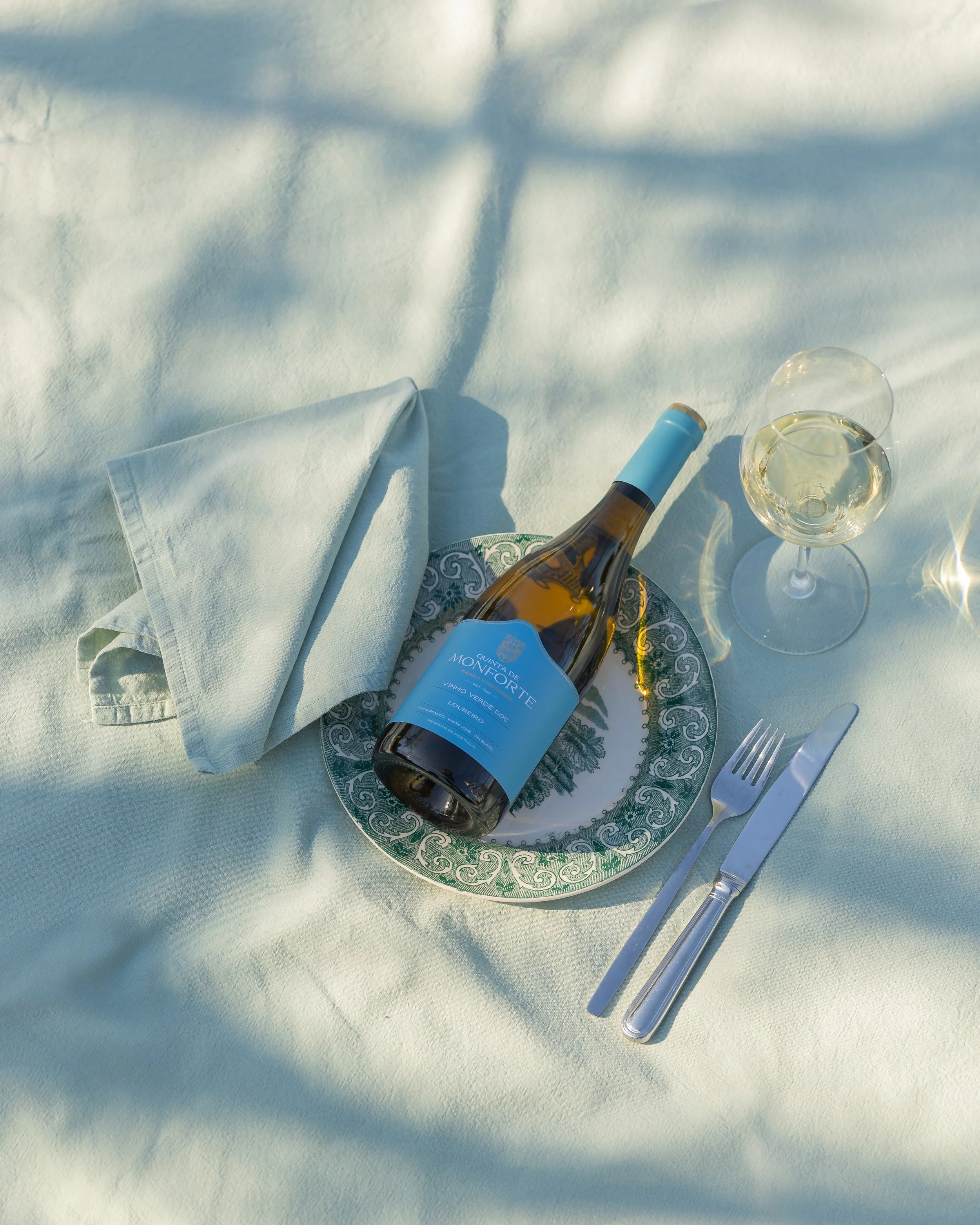 A bottle of white wine on a decorative plate with a green pattern, accompanied by a folded napkin, a glass of white wine, and a set of silver cutlery, all placed on a light blue cloth background.