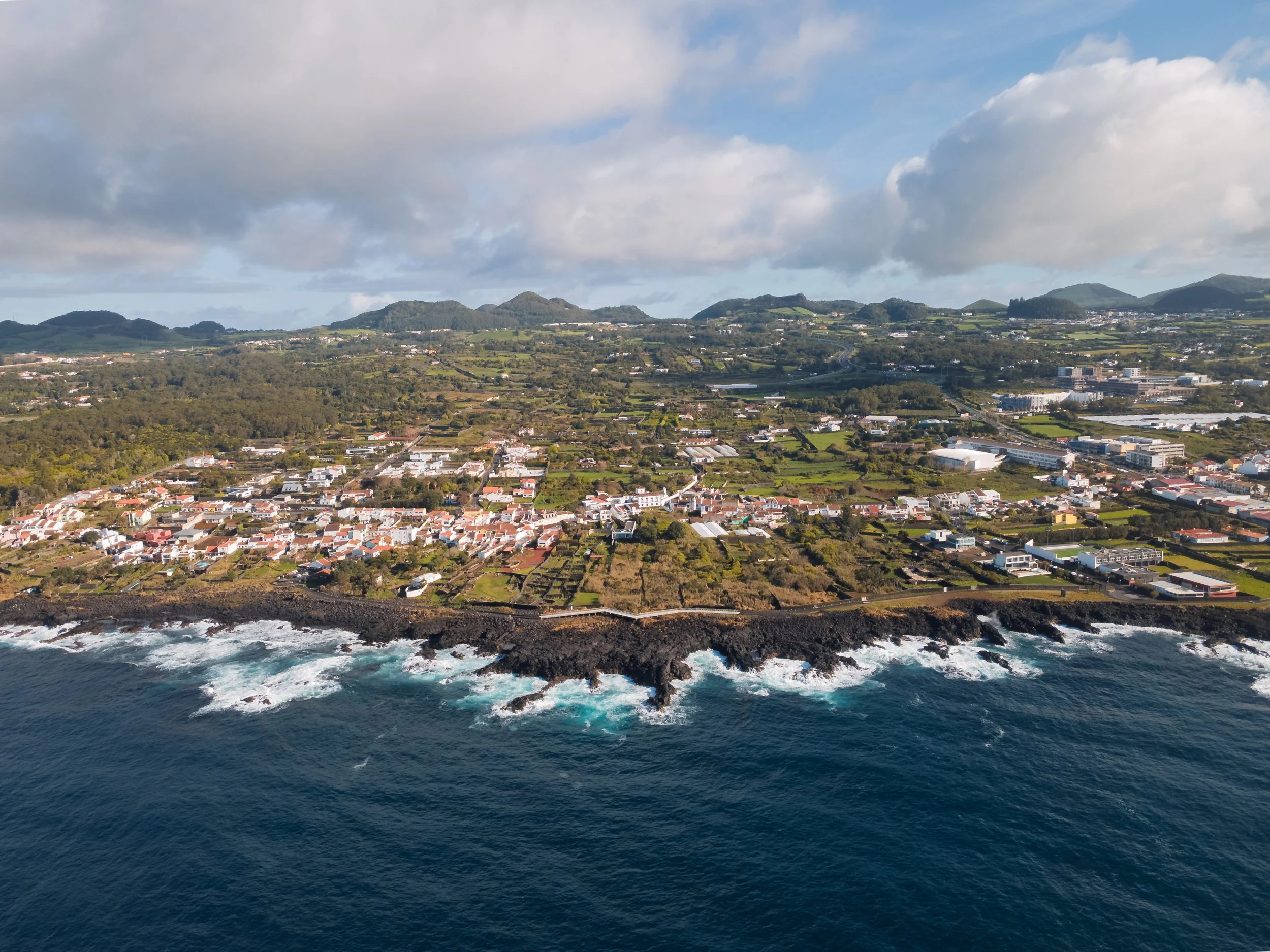 Aerial view of a coastal town with buildings and greenery, bordered by the ocean with rocky cliffs and waves crashing against them. In the background, there are hills under a partly cloudy sky.