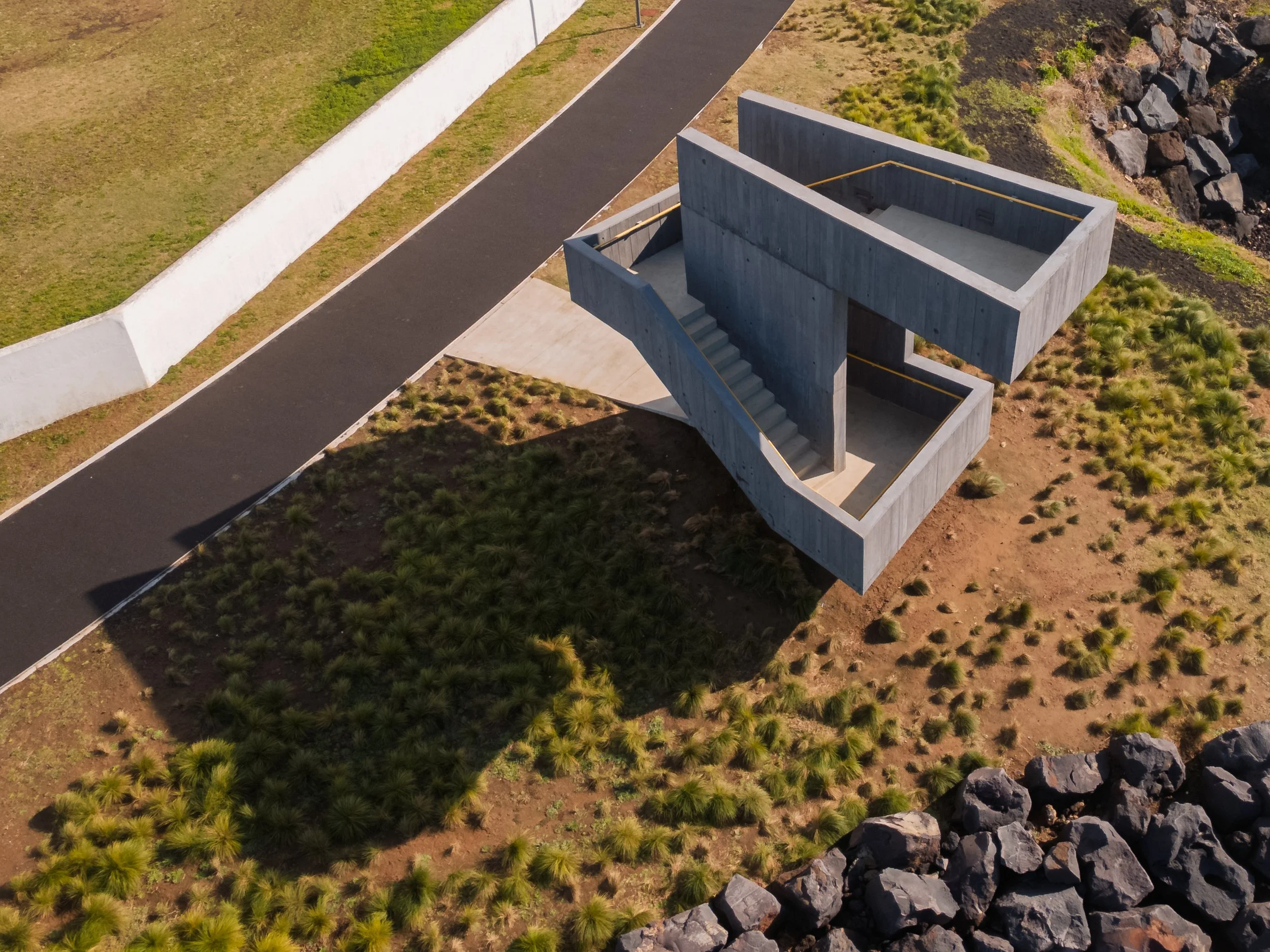 Aerial view of a modern, concrete, U-shaped staircase structure in a grassy landscape beside a paved pathway and rocky terrain.
