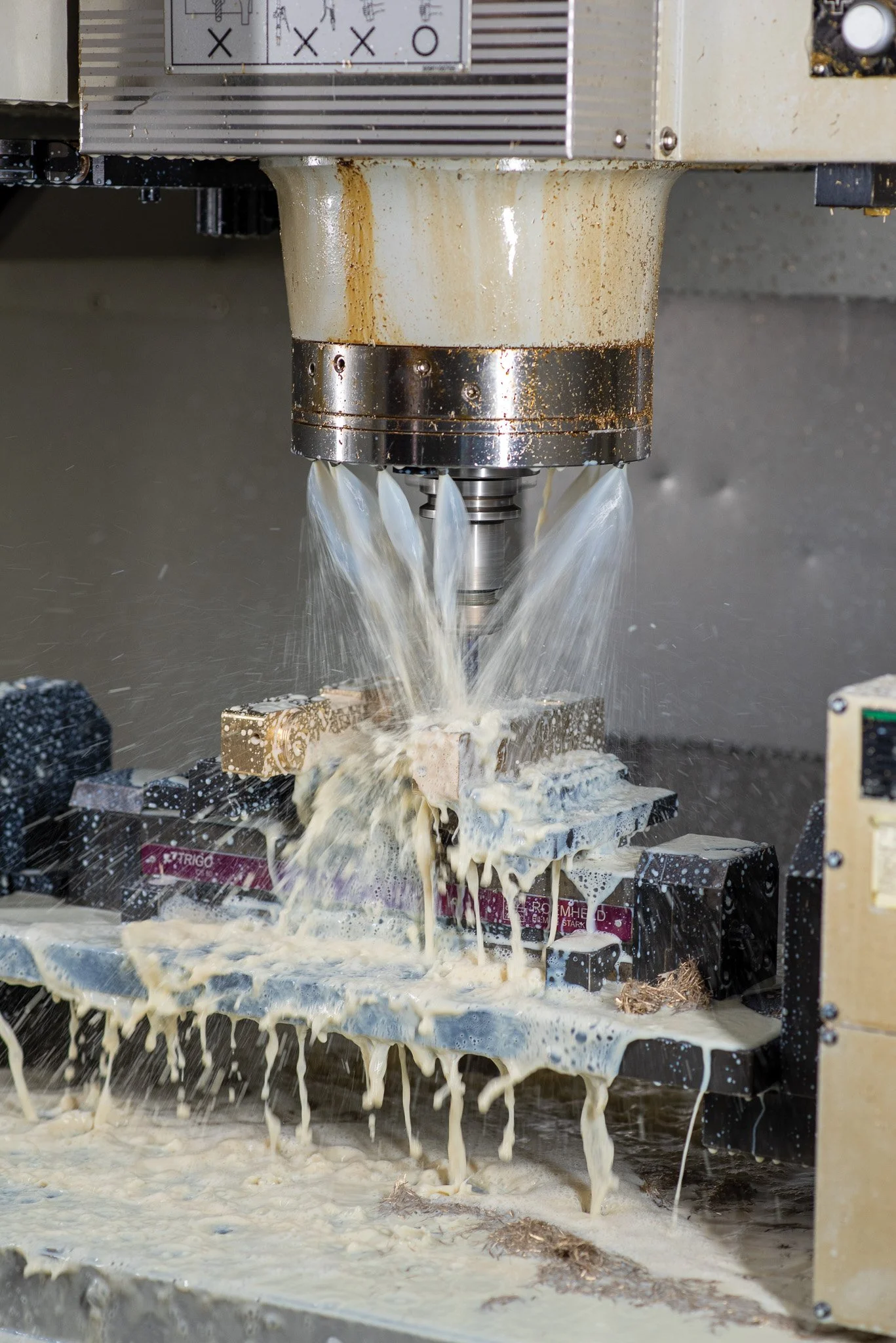 Máquina CNC em operação fabricando uma peça de metal, com líquido esguichando e espuma formando na superfície de trabalho.
