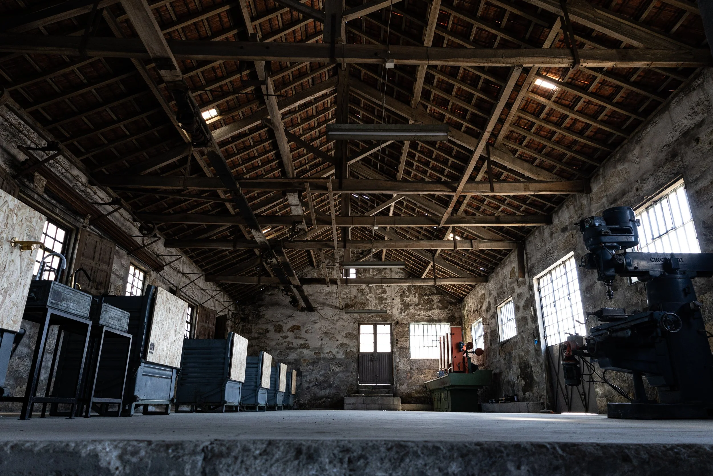 Interior de um galpão ou antiga fábrica com teto de madeira, janelas de vidro e equipamentos industriais, como uma máquina de perfuração. A luz natural entra pelas janelas e luzes de teto estão ligadas.