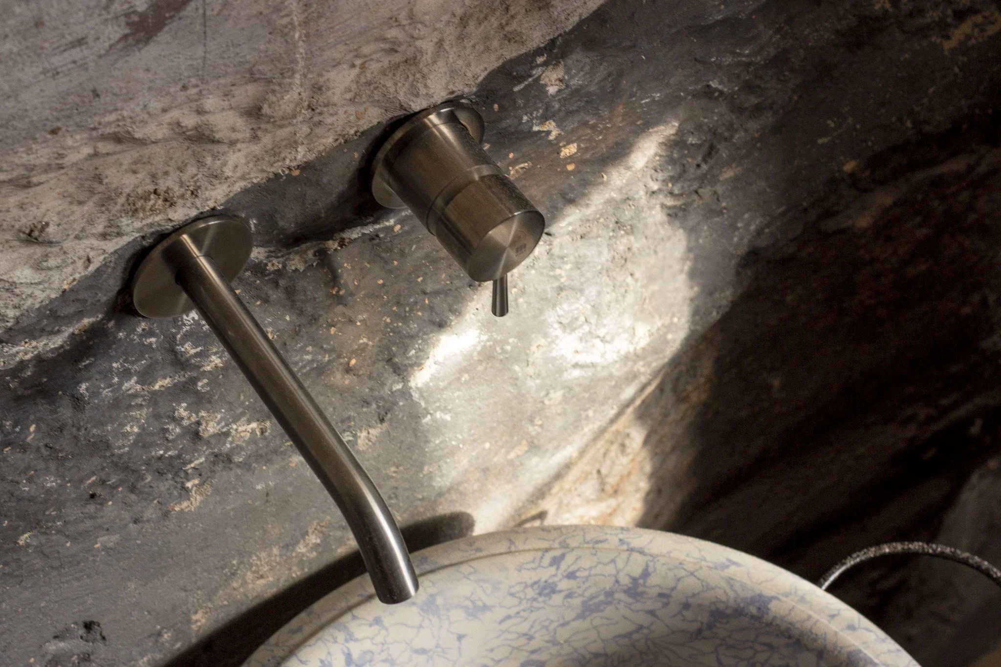Válvula de banho ou pia de metal instalada em uma parede de tijolos com revestimento de argamassa e cimento, ao lado de uma pia de cerâmica com superficie branca com desenhos azuis.