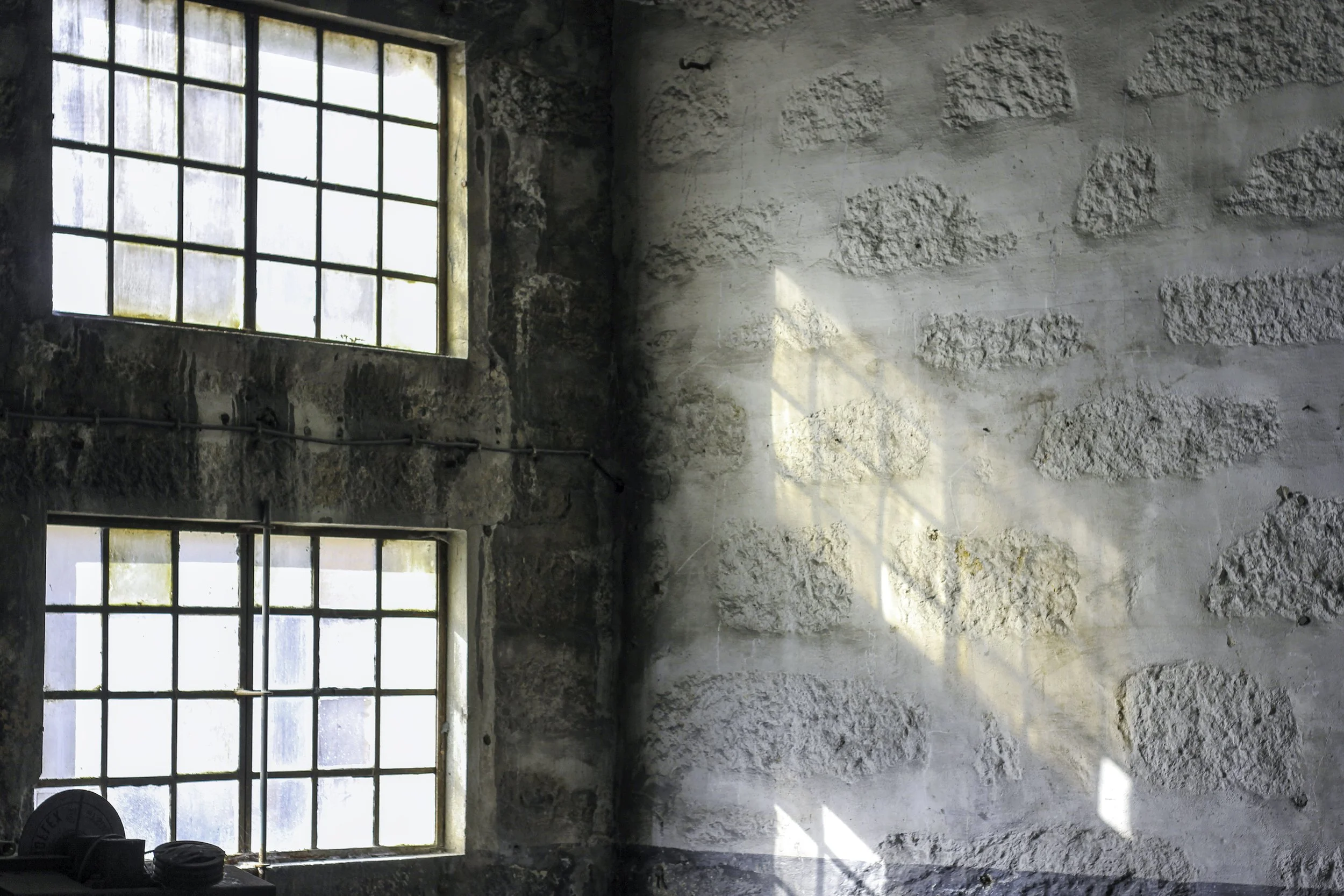 Janela grande de ferro com vidro antigo, parede de pedra e cimento, chão de concreto em uma sala vazia com equipamentos de academia no canto esquerdo.