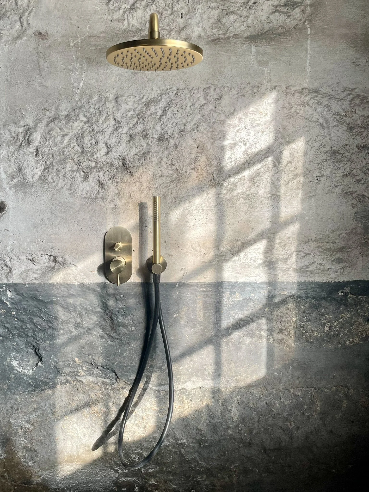 Chuveiro de teto dourado, com detalhes de controle na parede de pedra cinza, com cabo de ducha preto