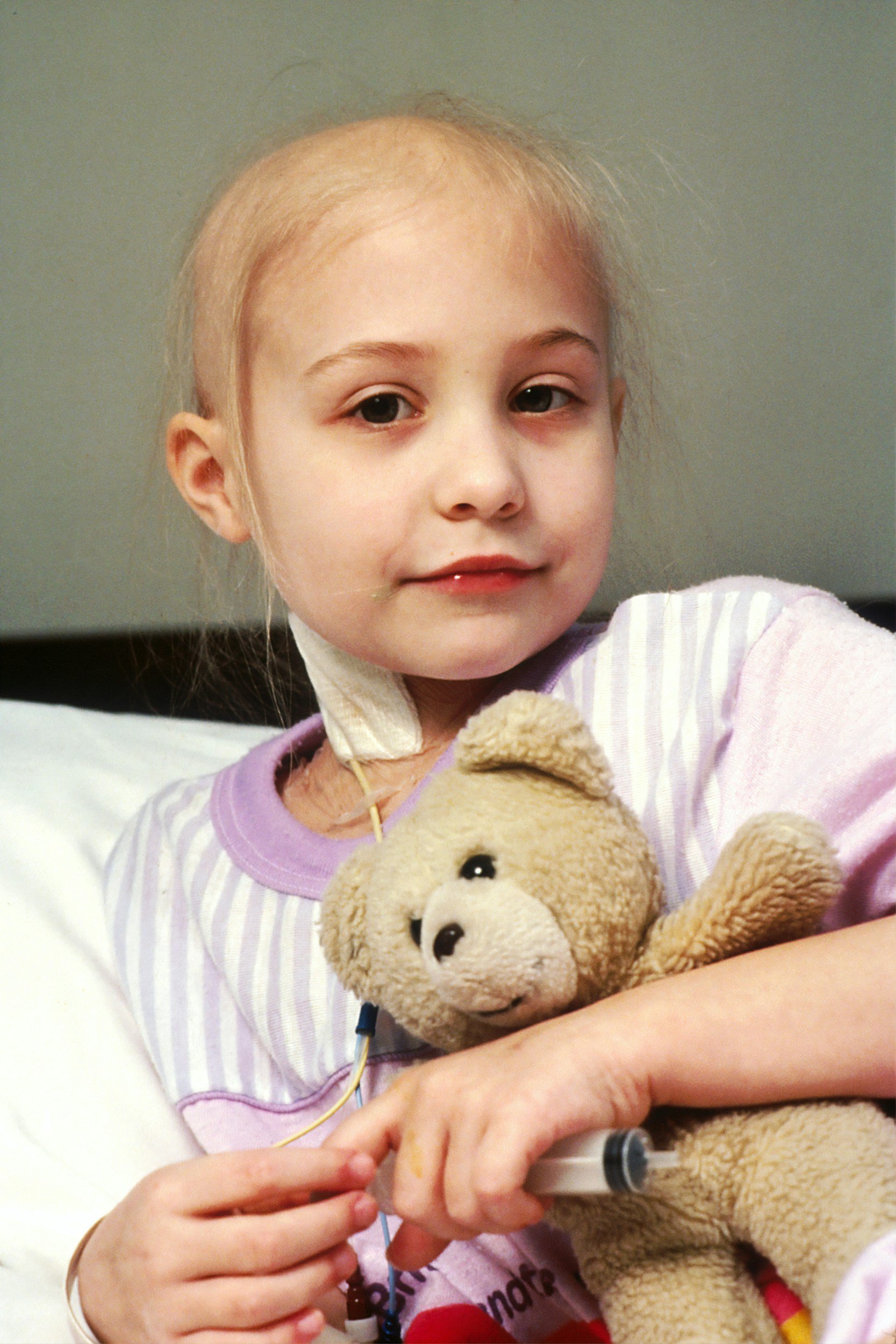 A young girl with blonde hair in a hospital bed holding a teddy bear in one hand and an IV in the other.