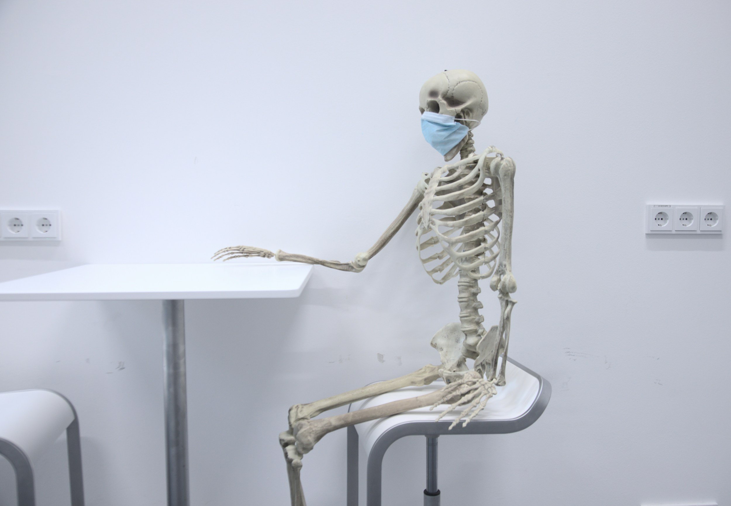 A human skeleton wearing a blue face mask, sitting on a chair in a plain white room with white walls, electrical outlets, and a white table.