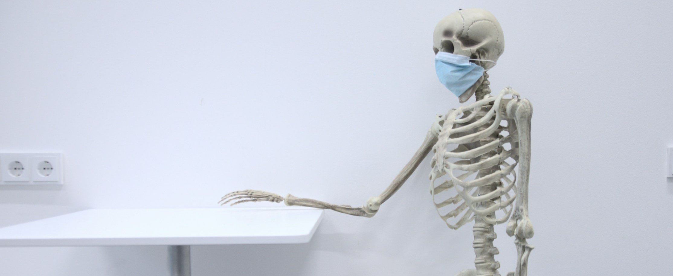 Skeleton wearing a blue face mask, sitting with one arm resting on a white table in a stark, white room.