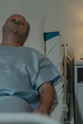 A man in a hospital bed with a medical gown, lying on his back, with a monitor and medical equipment beside him.