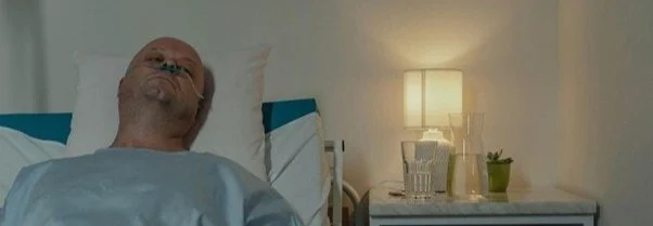 An elderly man lying in bed with eyes closed in a hospital, illuminated by a bedside lamp, with a glass of water and a small potted plant on a bedside table.