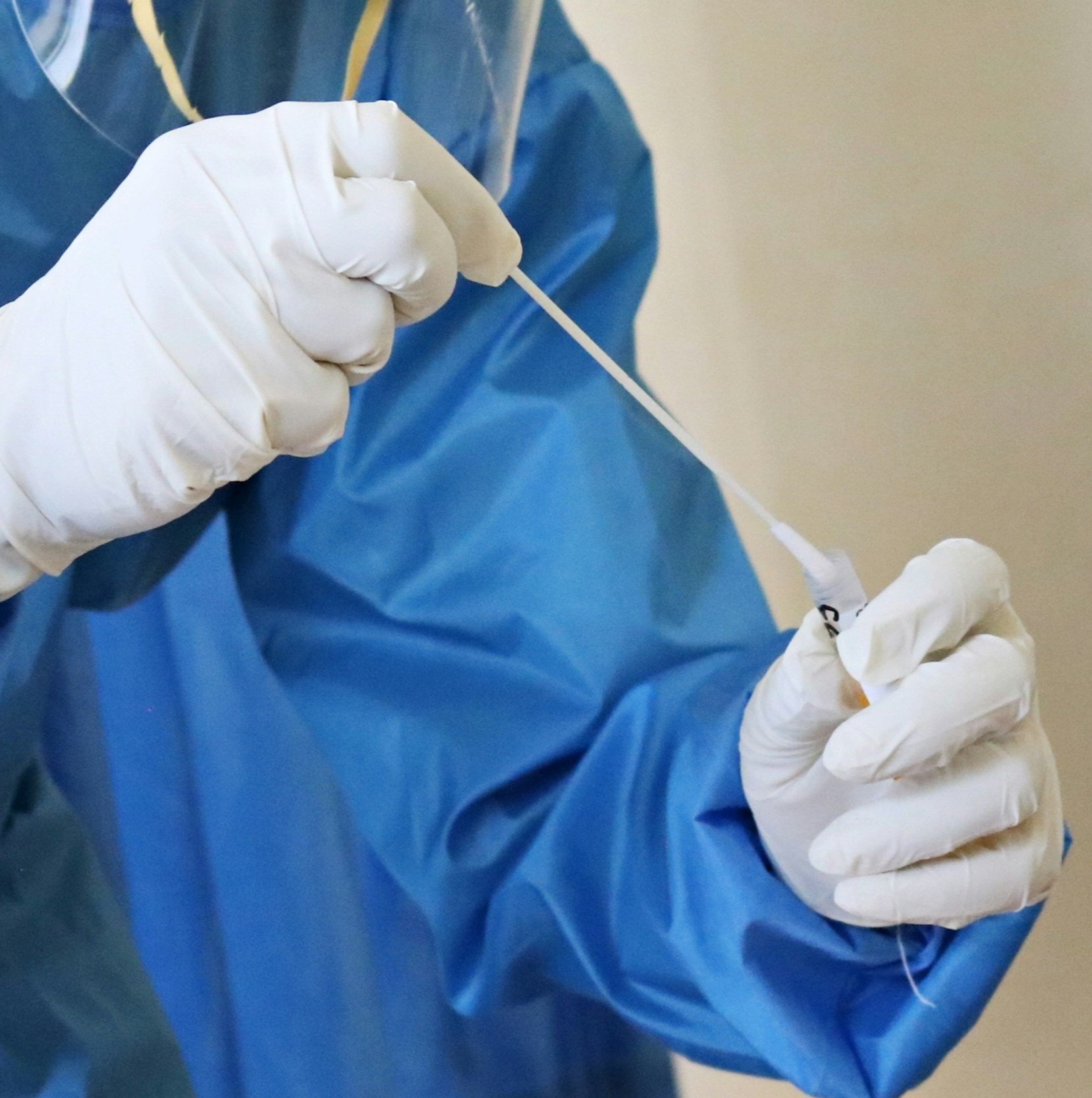 A healthcare worker wearing a blue gown and white gloves preparing a medical swab or test device.