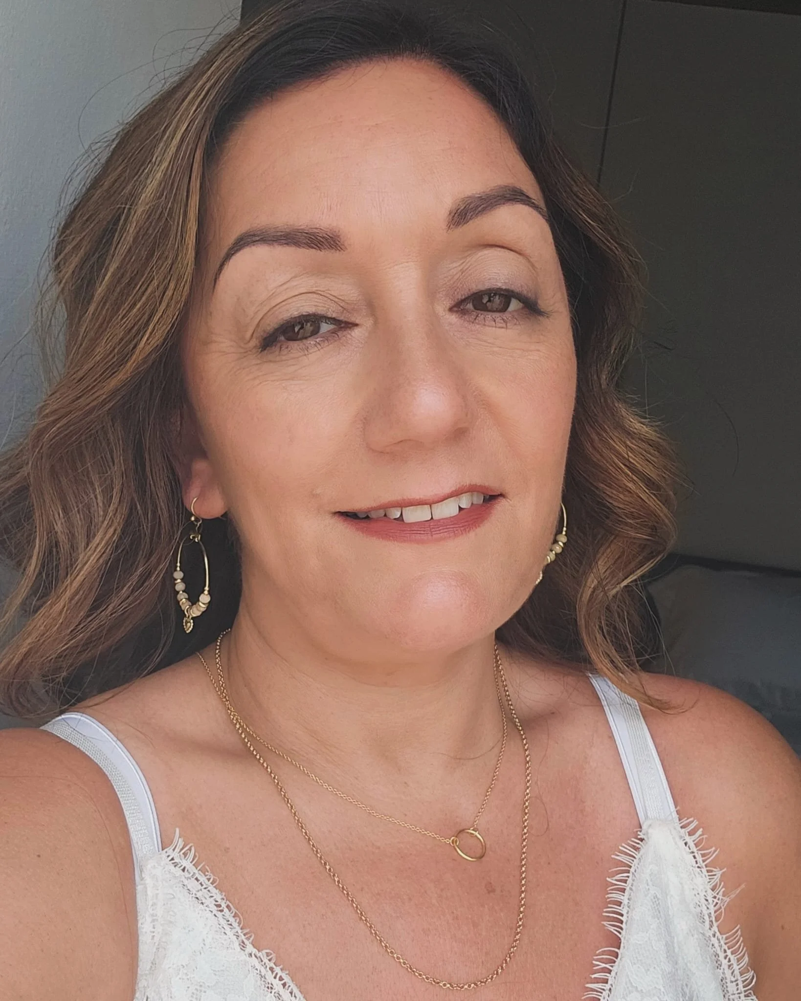 Close-up of a woman with light brown hair, wearing earrings and gold layered necklaces, smiling slightly, dressed in a white lacy top.