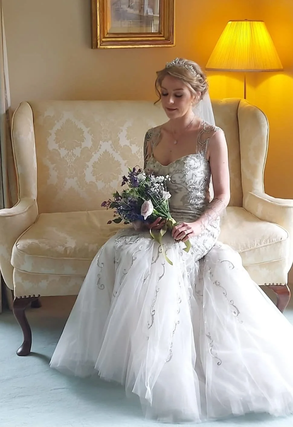 A woman in a wedding dress sitting on a vintage sofa, holding a bouquet of flowers, with a warm yellow lamp behind her.