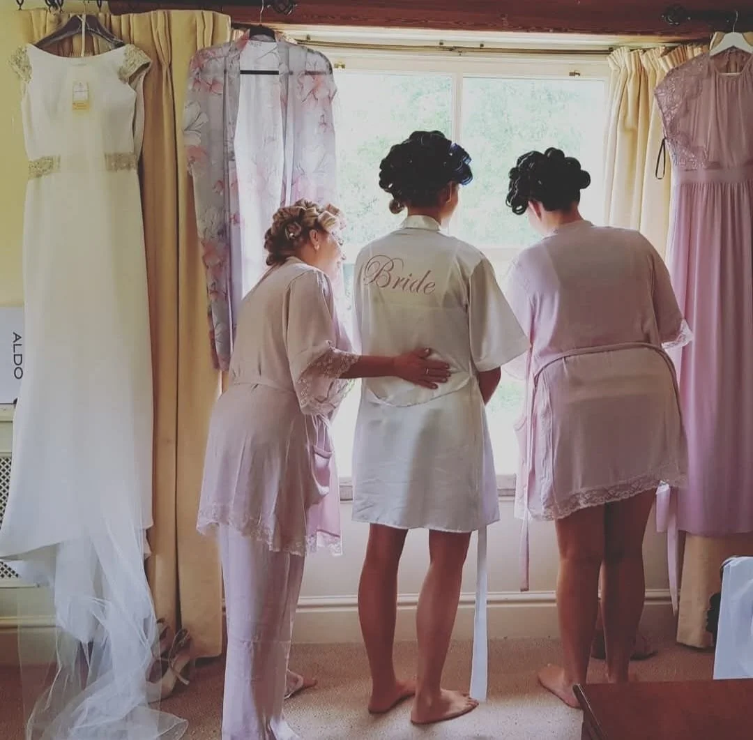 Three women, including a bride, preparing for a wedding, looking out a window in a room with hanging wedding dresses on either side. One woman has