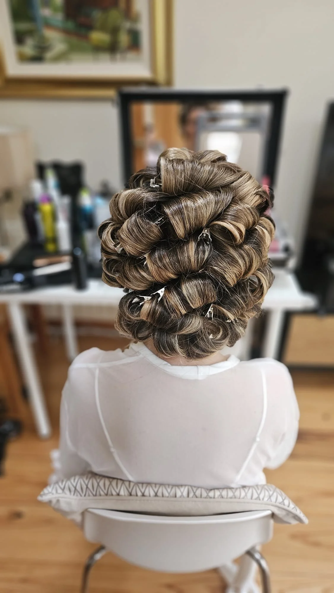 A person with a bathroom towel draped over their shoulders is sitting in front of a mirror, getting their hair styled into large, curled rollers with clips.