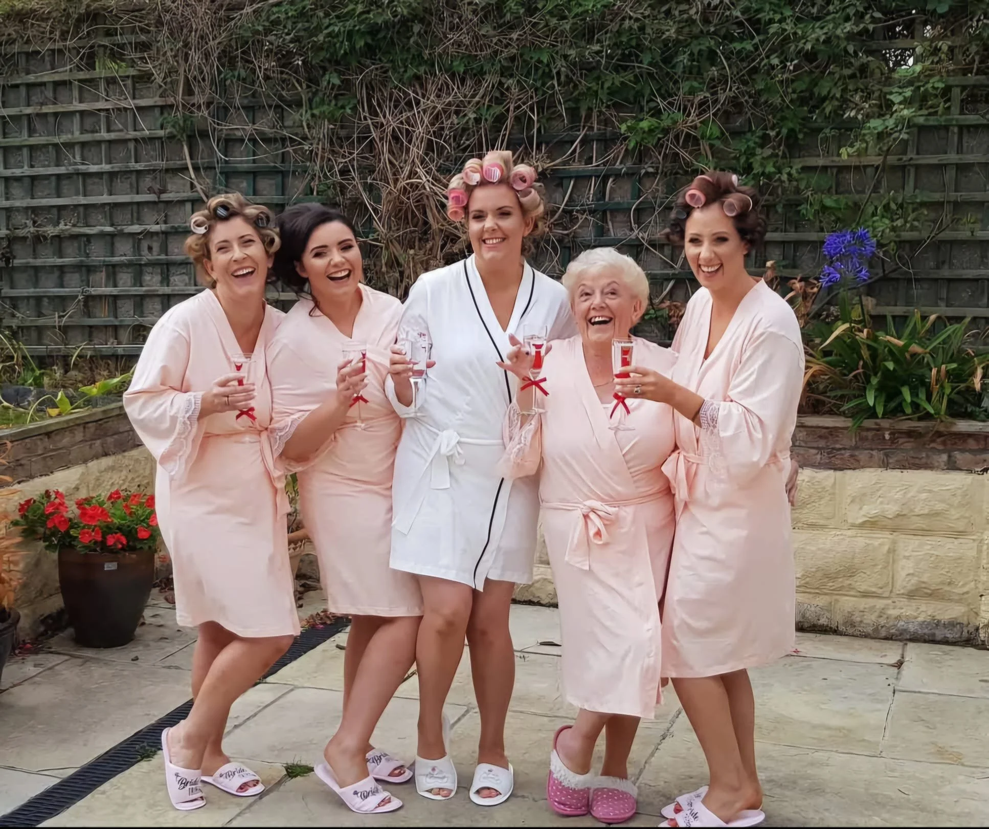 A group of six women, with five of them wearing pink nightrobes and one in a white robe, standing outdoors on a patio. They are all smiling, holding glasses with a red-colored drink, and appear to be celebrating. The women have their hair in curlers