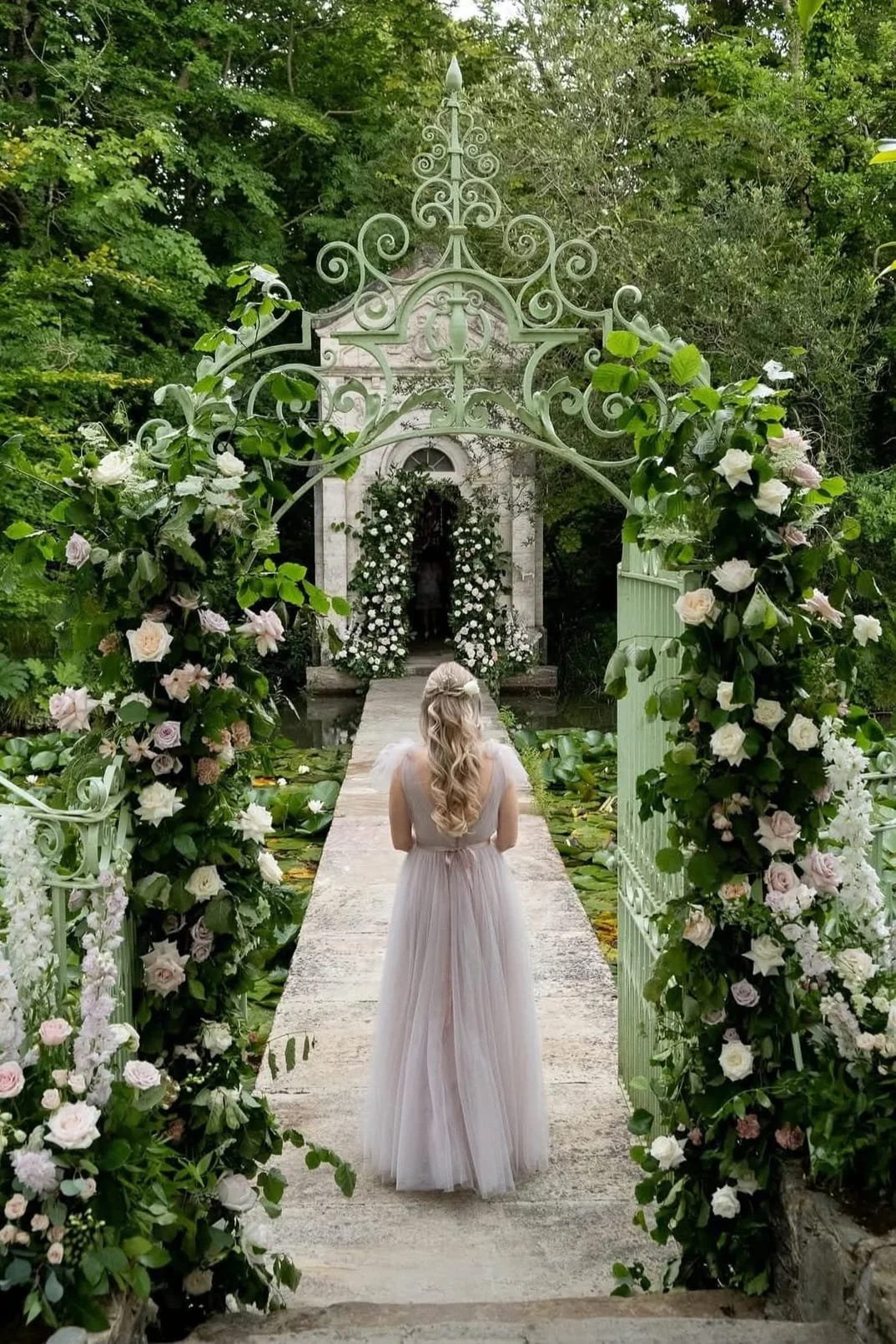 A woman with long, wavy blonde hair in a flowing, light pink dress standing on a stone path, facing a small, ornate white chapel decorated with flowers, surrounded by lush green trees.