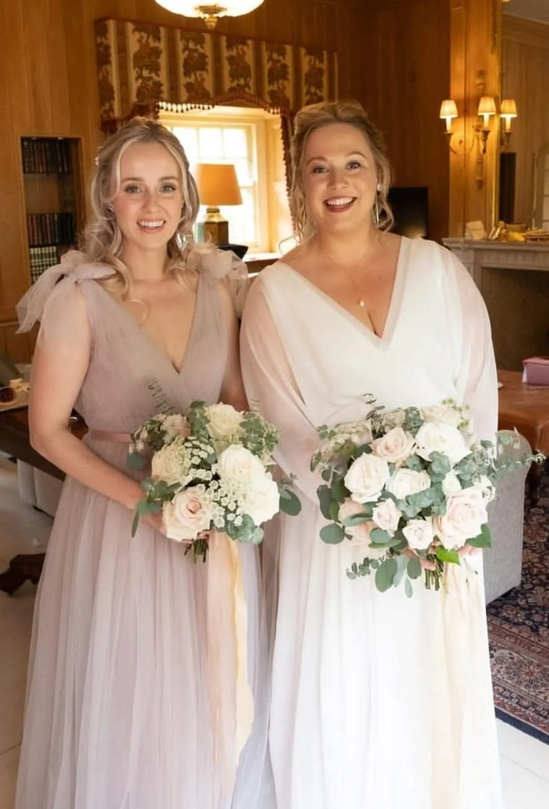 Two women in cream-colored dresses holding bouquets of white and blush roses and greenery, indoors with wooden walls and warm lighting.