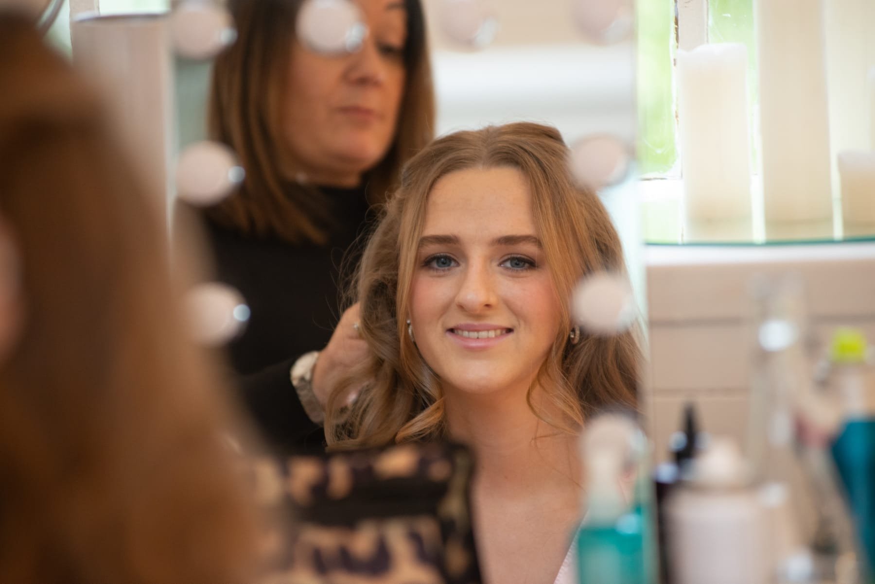 A young woman with long, curly auburn hair getting her hair styled in a mirror, with a stylist working behind her in a salon.