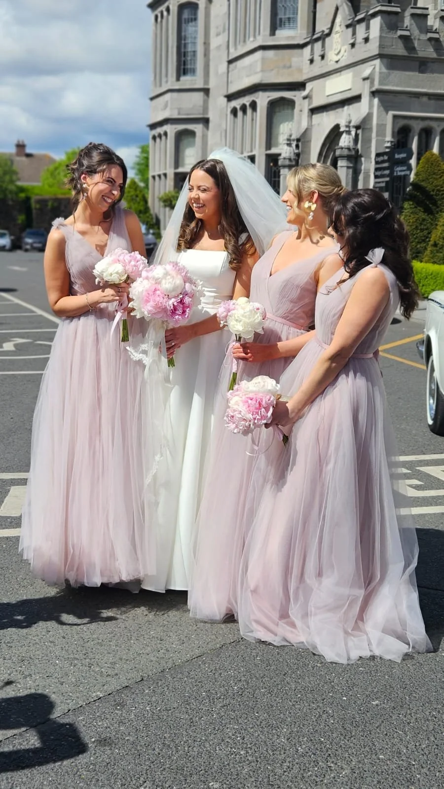 A bride in a white wedding dress holding a bouquet, smiling with four bridesmaids in light pink dresses holding pink and white bouquets, standing outdoors in front of a historic building.
