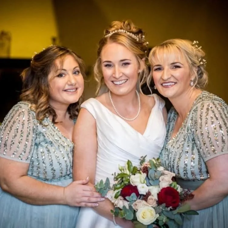Three women smiling, with one in a wedding dress holding a bouquet, and two women in matching gowns, at a wedding celebration.
