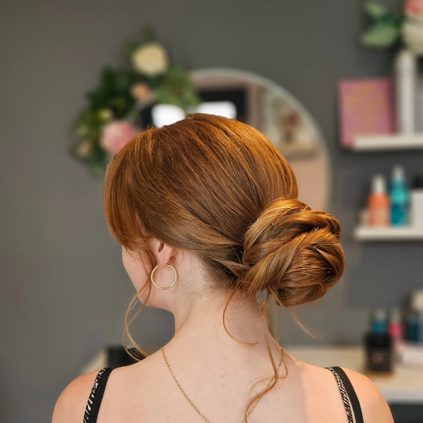 Back view of a woman with curly red hair styled in an elegant low bun, wearing gold hoop earrings and a black strap top, in a salon with blurred background.