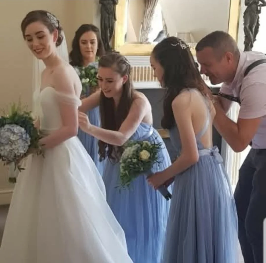 A bride in a white wedding dress is surrounded by bridesmaids in blue dresses, one of whom is adjusting the bride's dress while another holds a bouquet. A man is standing beside one of the bridesmaids, assisting or observing. The setting appears to b