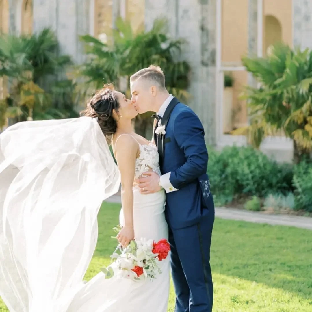 A bride and groom kiss outdoors on their wedding day. The bride wears a white dress with a veil, and the groom wears a blue tuxedo. The bride holds a bouquet of red and white flowers. There are green plants in the background.